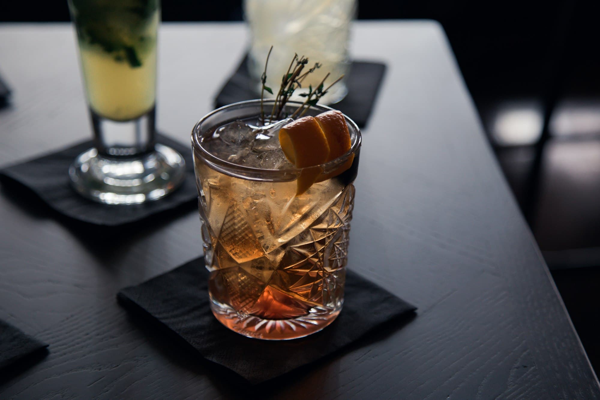 Old fashioned con scorza d'arancia e rametto di timo su un tavolo scuro, con altri cocktail sullo sfondo