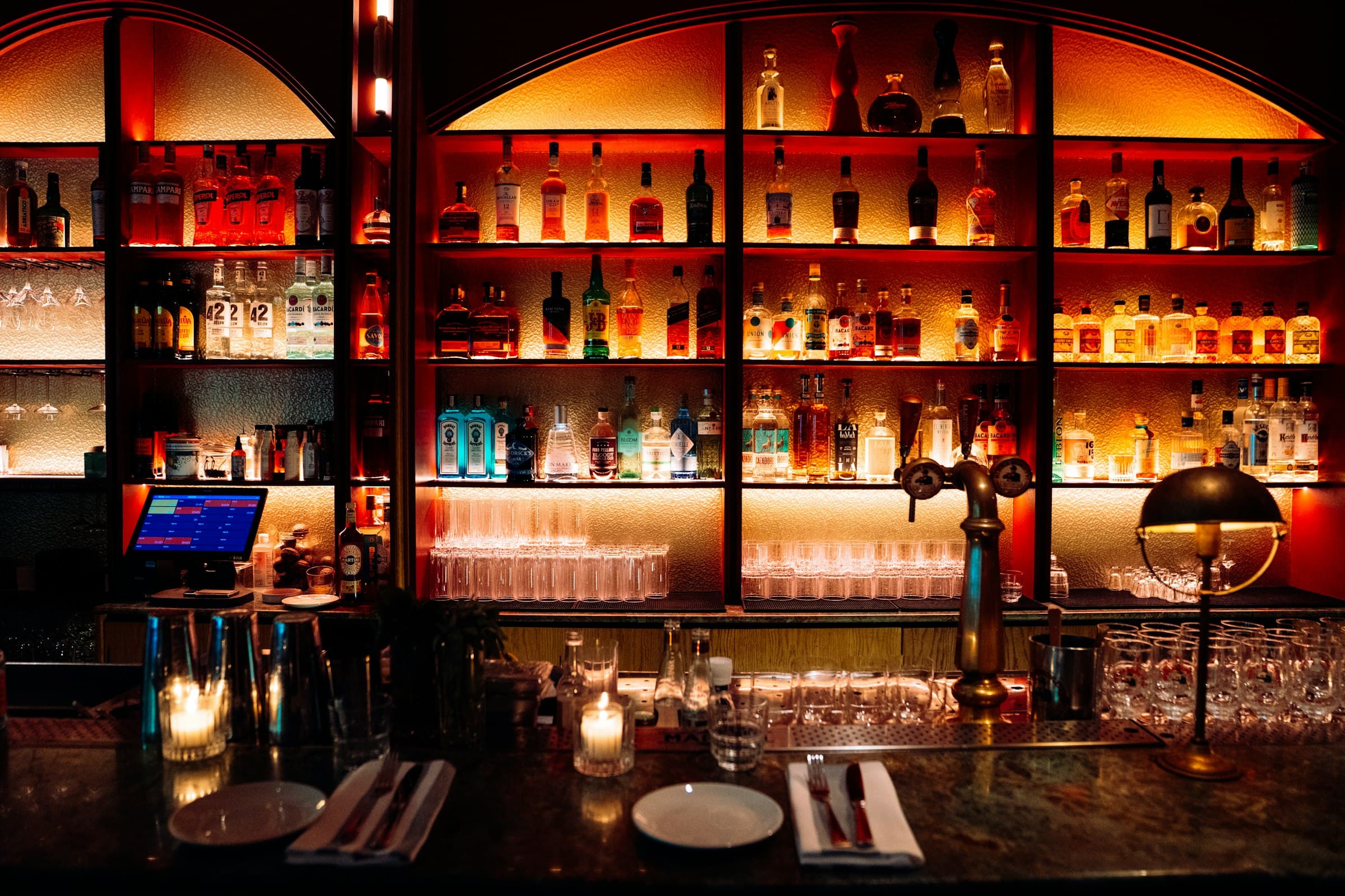 Parete di distillati retroilluminata dietro il bancone di Notte 21, con luci calde ambrate e rosse